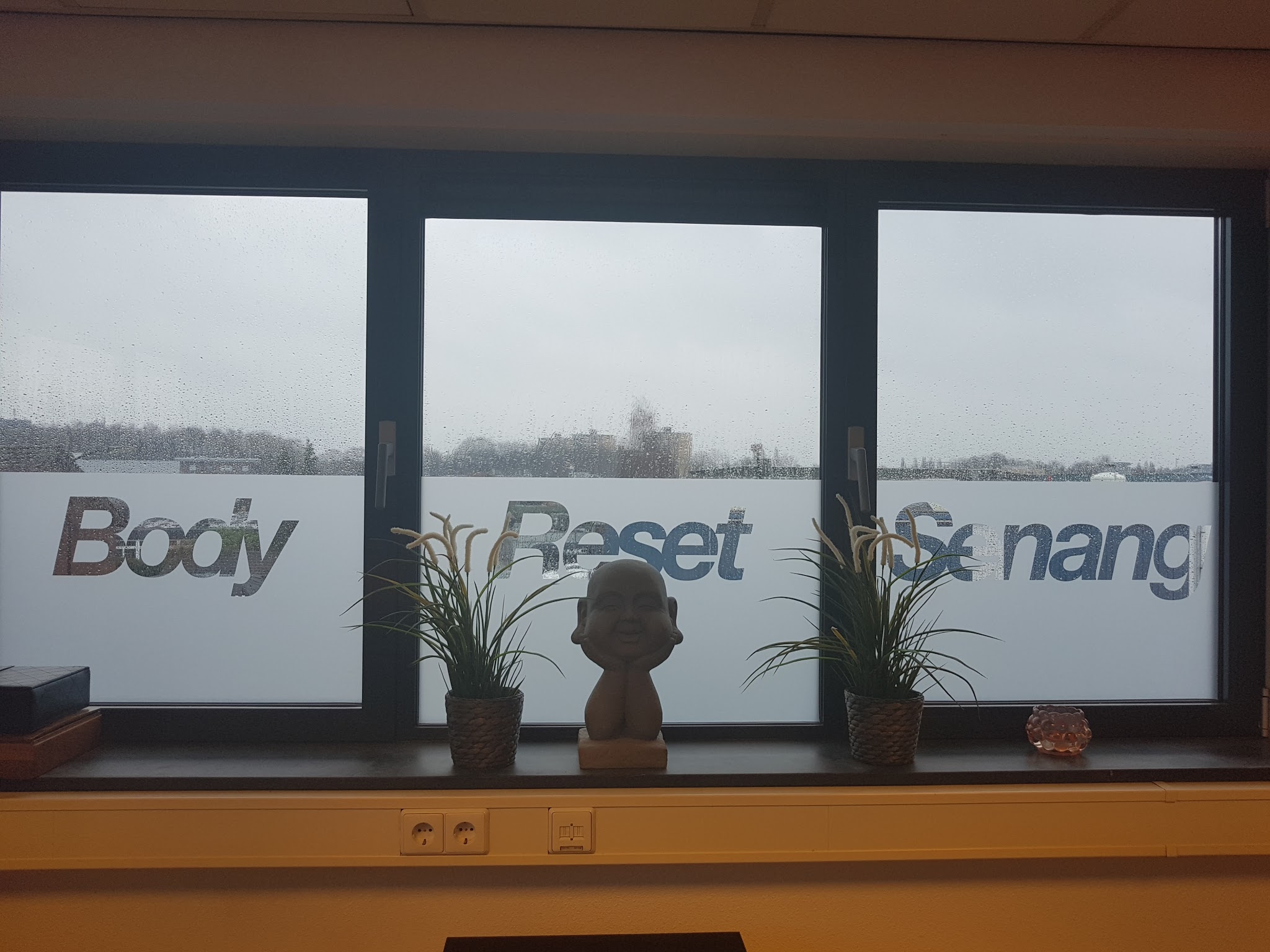 Het raam van de praktijk met de belettering Body Reset Senang en planten op de vensterbank